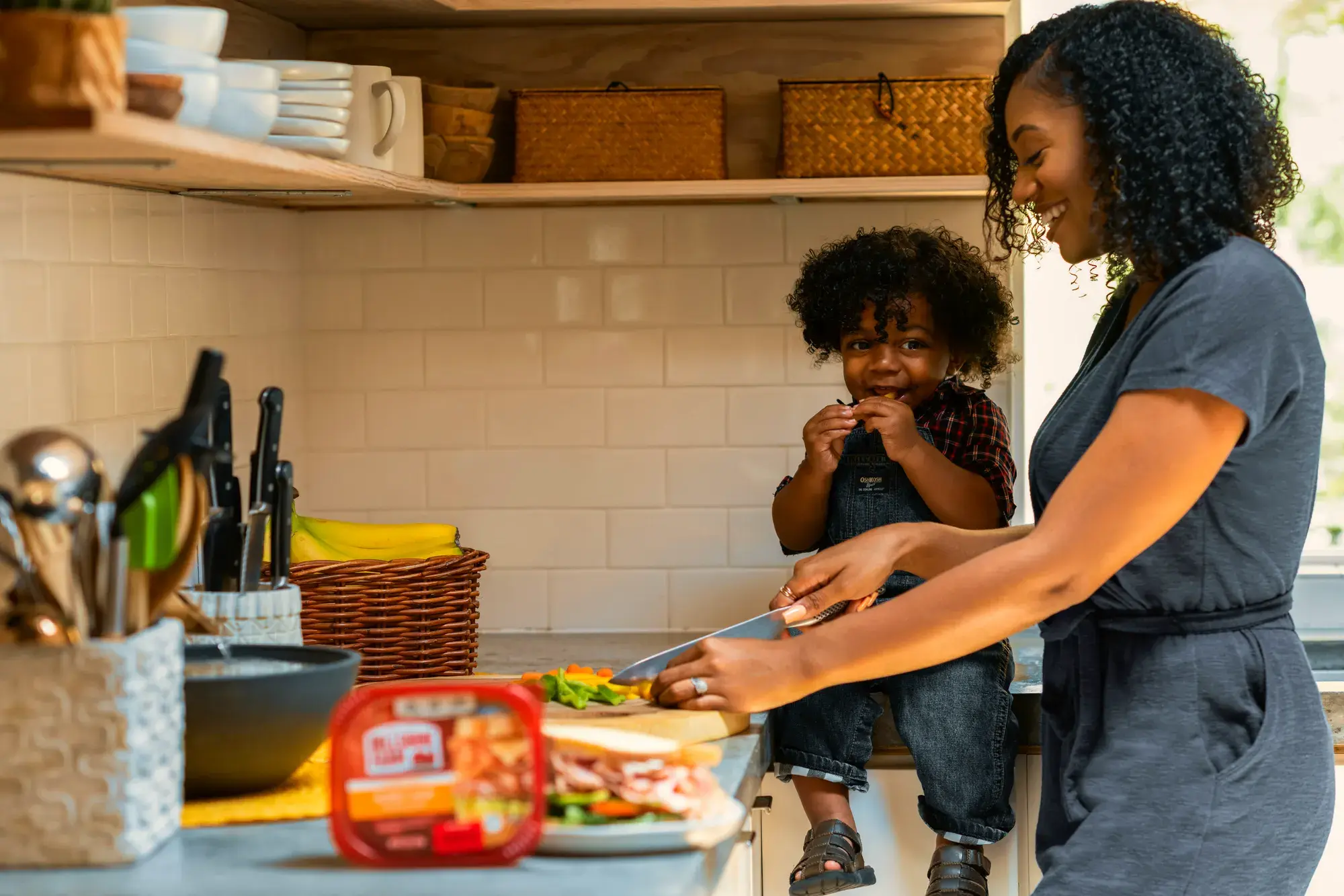 A parent preparing lunch with a small child sitting on the kitchen worktop